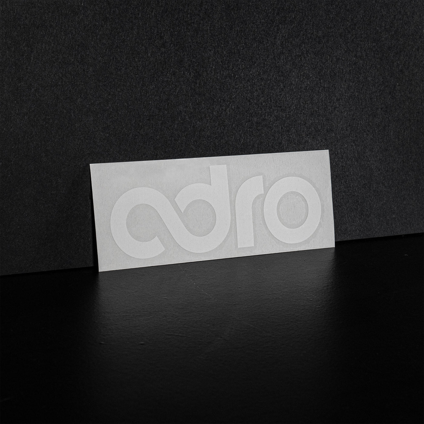 ADRO logo decal on a white rectangle with embossed adro lettering, detail view, carbon fiber-inspired background.