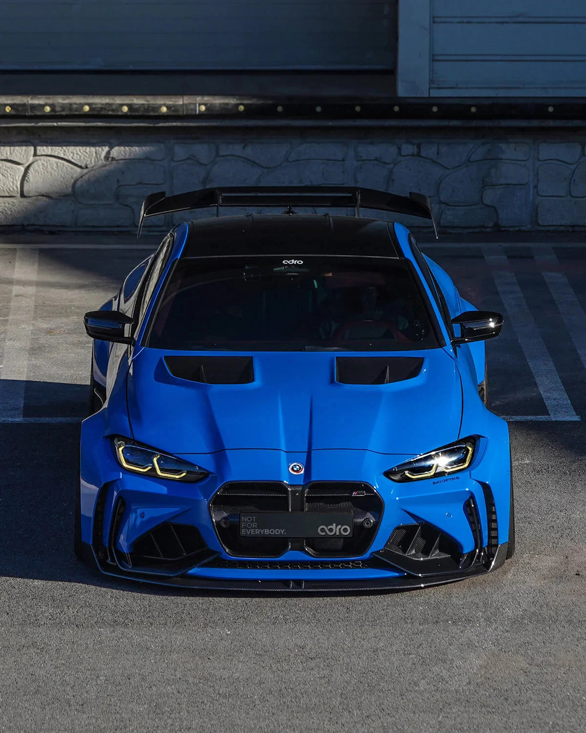 ADRO carbon rear wing on BMW G82 M4, rear three-quarter view, installed on blue car parked in front of building.