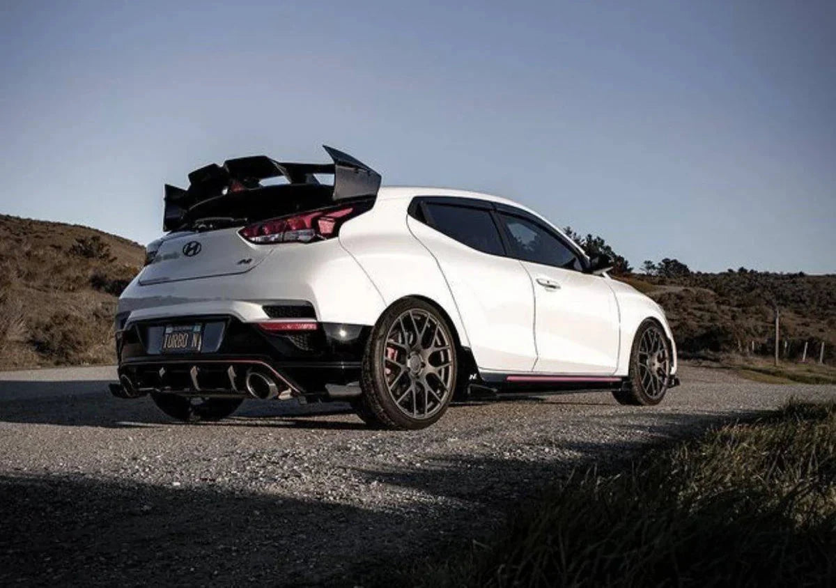 ADRO carbon fiber aero kit on Hyundai Veloster N, rear three-quarter view showing large spoiler, parked on gravel road.
